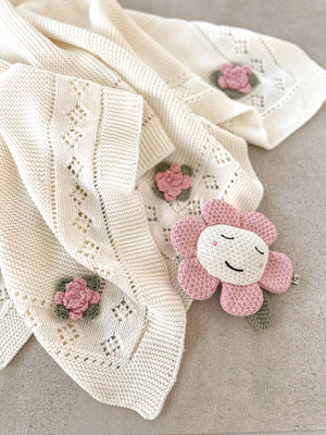 Cream knit blanket with pink crochet flowers and a smiling pink plush flower toy.
