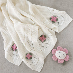 Cream knit blanket with pink floral patterns and a smiling pink flower plush toy beside it.