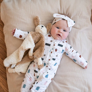 Infant in gift-patterned onesie and bow headband with plush bunny on beige blanket.