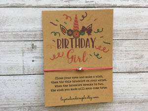 Brown card with "Birthday Girl" in colorful fonts, unicorn horn, pink thread bracelet, white wood background.