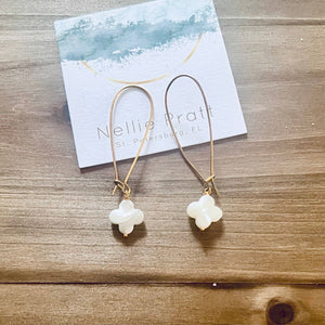 Gold hoop earrings with white clover pendants on a wooden surface, card in background.
