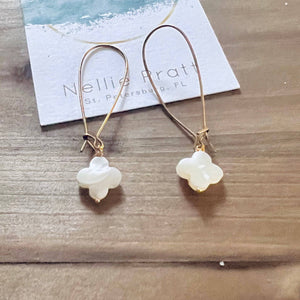 Gold hoop earrings with white clover charms on wood surface, blue and white card in background.