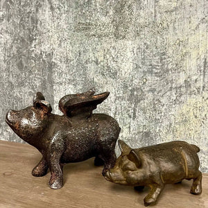 Two rustic metal pig sculptures, one with wings, on a wooden surface against a textured wall.