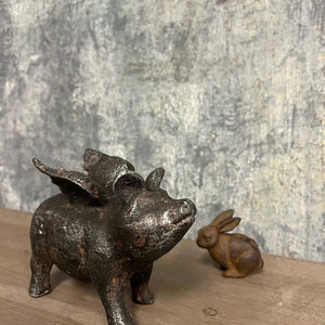Bronze statues of a winged pig and a rabbit on a wooden surface against a textured gray wall.