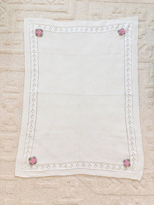 Cream blanket with lace border and pink floral crochet accents at each corner on a textured surface.