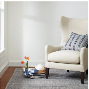 Cream armchair, blue-striped cushion, orange tulips in vase, woven tray with teapot on gray rug.