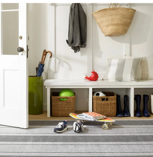 Entryway with baskets, a skateboard, shoes, and a striped rug. A coat, bag, and umbrellas are hung.