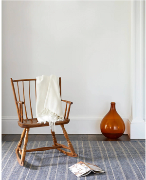 Wooden rocking chair with white blanket, amber vase, on blue-striped rug, against a white wall.
