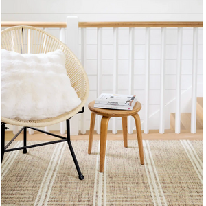 A woven chair with a white pillow next to a small wooden stool with books, on a striped beige rug.