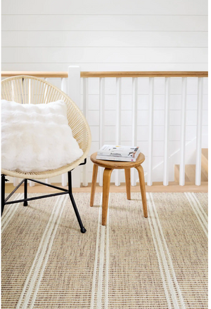A woven chair with a white pillow next to a small wooden stool with books, on a striped beige rug.