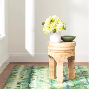 Wooden stool with white vase of yellow flowers and green bowl on green patterned rug.