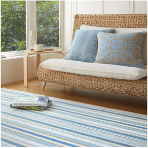 Woven sofa with blue patterned cushions, striped rug, small wooden table, and window with greenery.