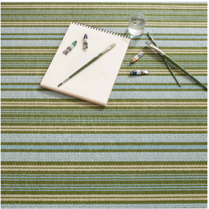 Notepad, brushes, and paint tubes on a green, blue, and beige striped surface with a glass of water.