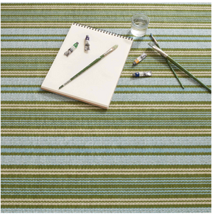 Notepad, brushes, and paint tubes on a green, blue, and beige striped surface with a glass of water.