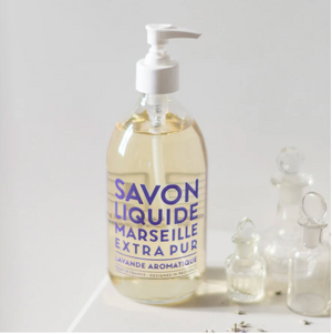 Clear soap bottle with purple text, white pump top, next to small clear glass bottles on white surface.
