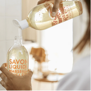 Hand pouring clear liquid soap from a plastic bottle into a glass bottle with orange text.
