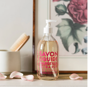 Clear soap bottle with pink text, on a shelf with rose petals, brush, and floral artwork.