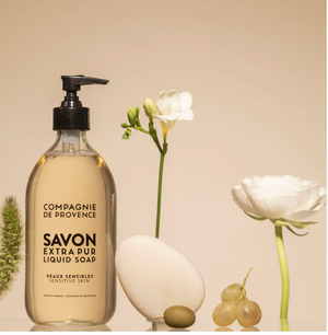 Clear soap bottle with pump, surrounded by white flowers, green grapes, and an olive on a beige background.