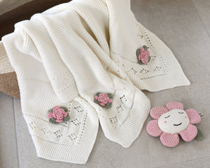 Cream knitted blanket with pink flower patterns, next to a smiling pink flower plush on gray tile.