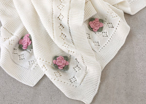 Cream knit blanket with pink flower patterns and diamond lace details, on a gray surface.