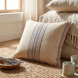 Beige pillow with blue stripes on a textured blanket, near a window and woven mat with cups and lavender.