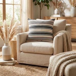 Beige armchair with striped pillow, knitted throw, pampas grass vase, and wooden dresser in sunlight.
