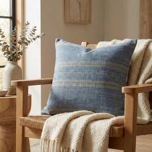 Blue striped pillow on a wooden chair with a cream knit throw, next to a vase with eucalyptus.