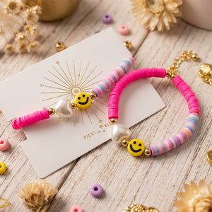 Colorful bracelets with pink and pastel beads, yellow smiley faces, and pearl hearts on wood.