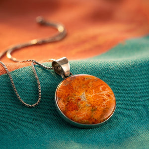 Round orange stone pendant on a silver chain, placed on a teal and orange fabric background.