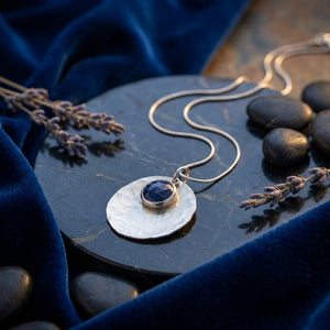 Silver necklace with blue gemstone pendant on dark marble, surrounded by lavender and velvet.
