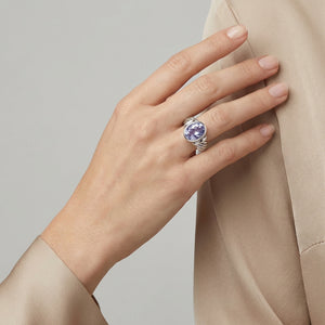 Hand wearing a silver ring with a large purple gemstone, set against a beige fabric background.