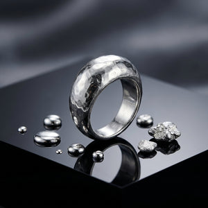 Silver ring and droplets on a glossy black surface, with scattered metallic pebbles.