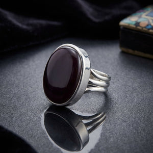 Silver ring with an oval dark stone on a reflective gray surface, with a black fabric background.
