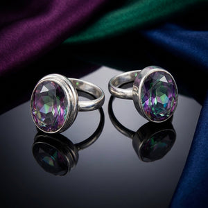 Two silver rings with oval, multicolored gemstones on a reflective black surface, draped fabric backdrop.