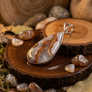 A teardrop agate pendant with brown and white stripes on wooden discs, surrounded by polished stones.