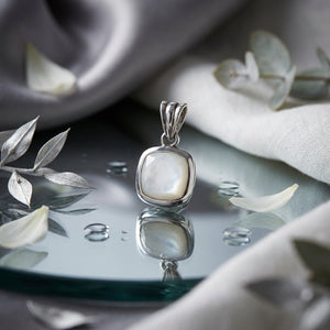 Silver pendant with pearl on reflective surface, surrounded by petals and soft gray fabric.