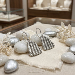 Silver textured earrings on beige cloth, surrounded by white and silver stones, dried flowers.