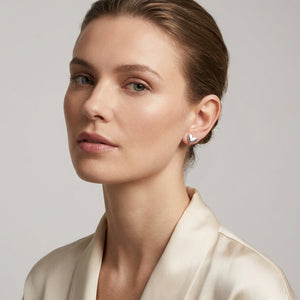 Woman with short hair wears silver heart earrings and a cream blouse against a gray background.