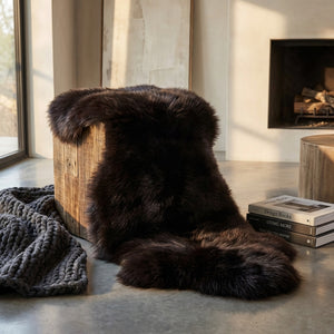 Dark fur rug draped over wooden stool, gray knit blanket, books nearby, modern interior setting.