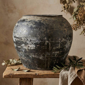 A weathered, dark ceramic pot on a wooden table with dried leaves and branches around it.