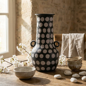 Tall black vase with white polka dots on a wooden table, surrounded by white flowers and bowls.
