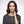 Woman with wavy brown hair, wearing a gray top and an orange pendant necklace, against a gray background.