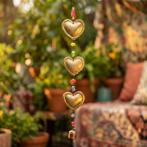 Golden heart ornaments with colorful beads hang vertically, with blurred plants in the background.