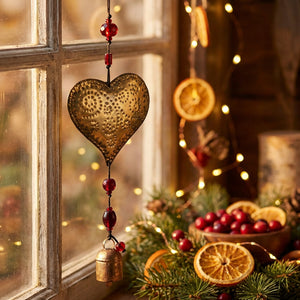 Golden heart ornament with red beads and bell by a window, surrounded by festive lights and greenery.
