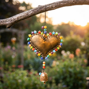 Bronze heart ornament with colorful beads and a bell, hanging from a branch at sunset.