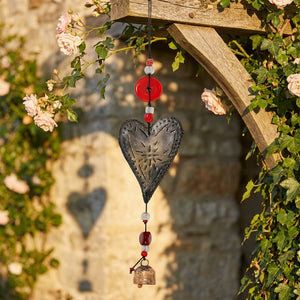 Metal heart wind chime with red beads, hanging on a wooden arch near pink roses and green leaves.