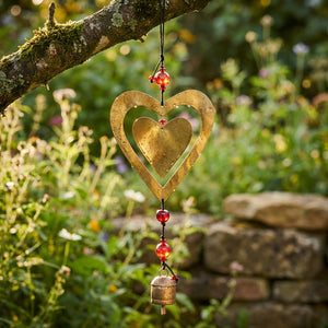 Golden heart wind chime with red beads hanging from a branch in a lush garden.