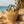 A woven beach bag with lemons, flowers, and a hat sits on sandy driftwood by the ocean.