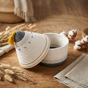 Woven basket with lid, white and gray patterns, yellow tassel, on wooden surface with cotton stems.
