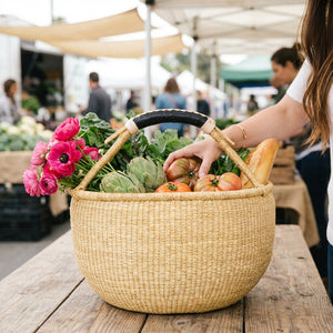 Woven basket on wooden table with pink flowers, green vegetables, tomatoes, and baguette.